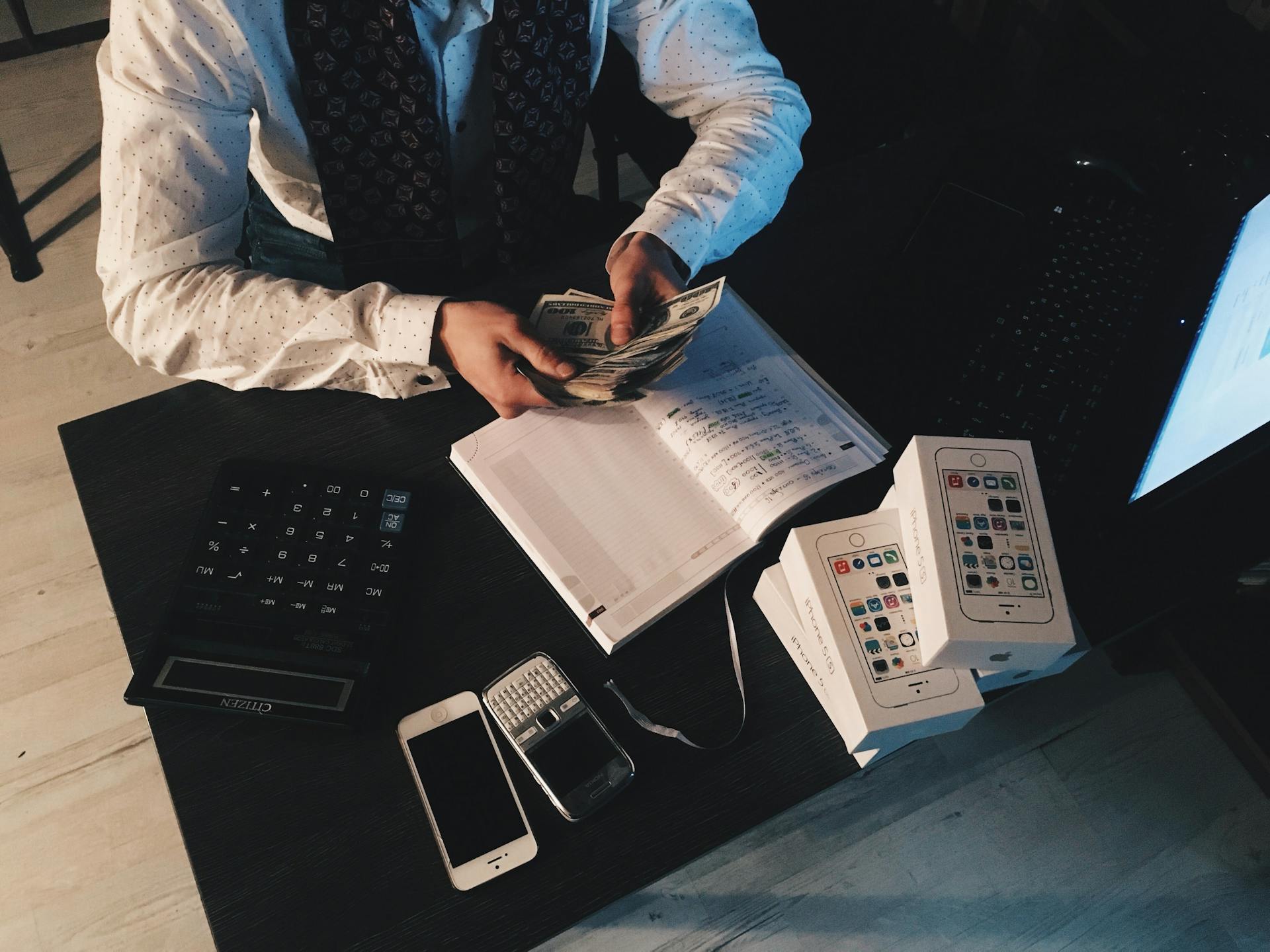 A man (bookkeeper) holding a large amount of US Dollars counting them while sat at a desk which had books, mobile phones and a calculator on it.